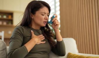 Junge Frau, die sich beim Atmen schwer tut und einen Inhalator benutzt, während sie ihre Brust hält und Asthmasymptome und Atembeschwerden zeigt.