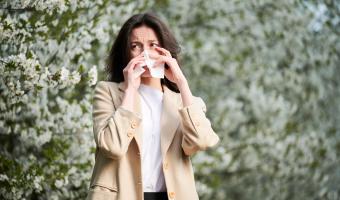 Woman allergic suffering from seasonal allergy at spring in blossoming garden at springtime. Young woman sneezing and blowing nose with nasal handkerchief in front of blooming tree. Allergy concept.