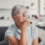 Sad elderly woman sits alone on a sofa at home, looking out the window with a worried expression. She reflects on challenges and feels stressed, illustrating loneliness and concern.